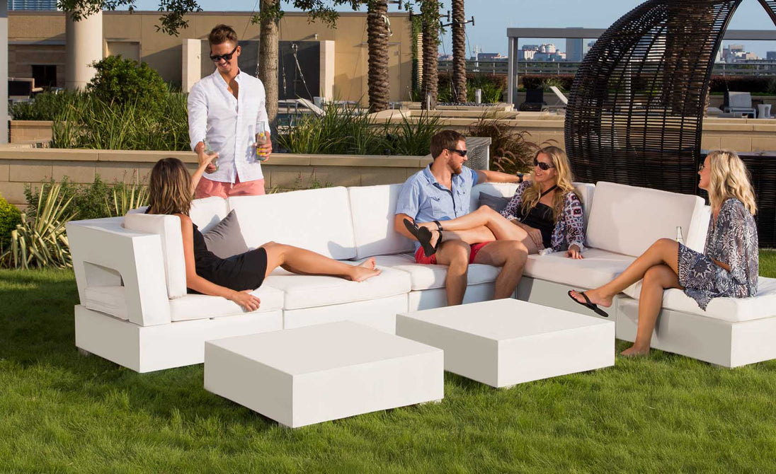 Four people relax in a garden setting with Signature Sectional Endcap couches by Ledge Lounger. One stands with a drink near the ottoman, while others are seated and chatting. Greenery blends with a cityscape backdrop, creating an inviting corner for their gathering.