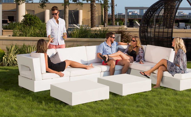 Four people relax in a garden setting with Signature Sectional Endcap couches by Ledge Lounger. One stands with a drink near the ottoman, while others are seated and chatting. Greenery blends with a cityscape backdrop, creating an inviting corner for their gathering.