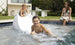 Two kids in swimsuits have fun on the Ledge Lounger Signature Slide, splashing into the pool and smiling. A woman in a floral dress and sunglasses relaxes by the tanning ledge, watching them. A building and greenery create a peaceful backdrop.