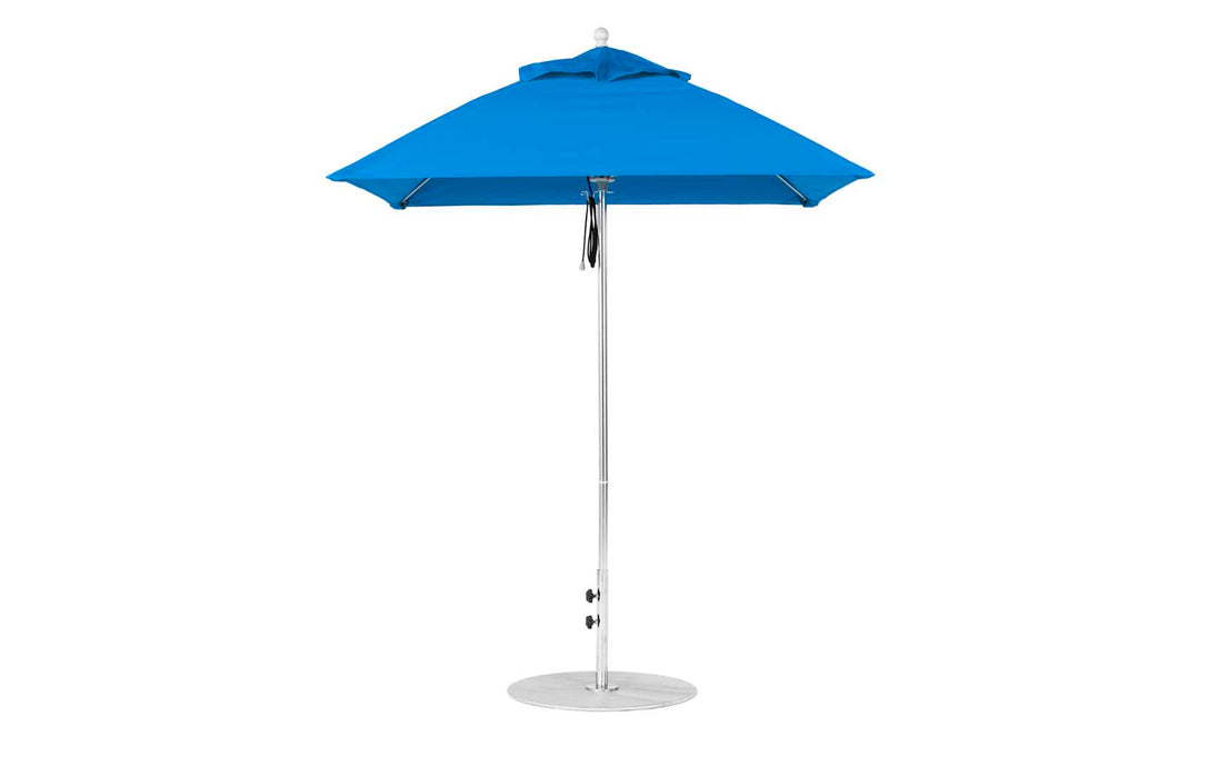 The Essential Fiberglass Umbrella by Ledge Lounger, a 6.5 square pulley model, has a blue canopy made of marine-grade fabric with an aluminum pole on a white round base. Its triangular shape offers shade and elegance, standing isolated on a white background.