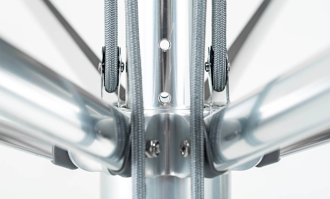 Close-up of the Premier Aluminum Umbrellas wind-resistant structure, featuring converging cylindrical rods at a central hub, secured with bolts and gray cables by Ledge Lounger; showcased on a plain white background.
