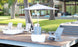 A modern house enhances the peace of a lakeside deck, featuring a PartyPad Swim-Up Bar by Ledge Lounger and Adirondack chairs around a low table. With a white parasol nearby, its an ideal spot for poolside sipping with your favorite drink.
