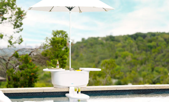 The PartyPad Swim-Up Bar by Ledge Lounger, featuring a small white table with a parasol, Corona beer, limes, and a shaker on top, creates the perfect poolside sipping setup against a backdrop of tranquil water and lush greenery.
