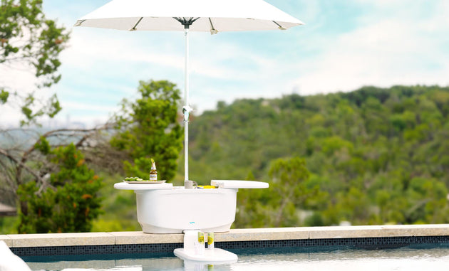 The PartyPad Swim-Up Bar by Ledge Lounger, featuring a small white table with a parasol, Corona beer, limes, and a shaker on top, creates the perfect poolside sipping setup against a backdrop of tranquil water and lush greenery.
