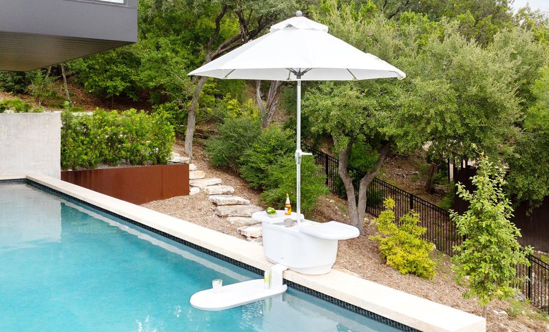 The PartyPad Swim-Up Bar by Ledge Lounger, with a white umbrella, is in the pool offering drinks and snacks. Surrounded by lush greenery, the serene outdoor area features stone and metal fencing, enhancing a tranquil atmosphere.