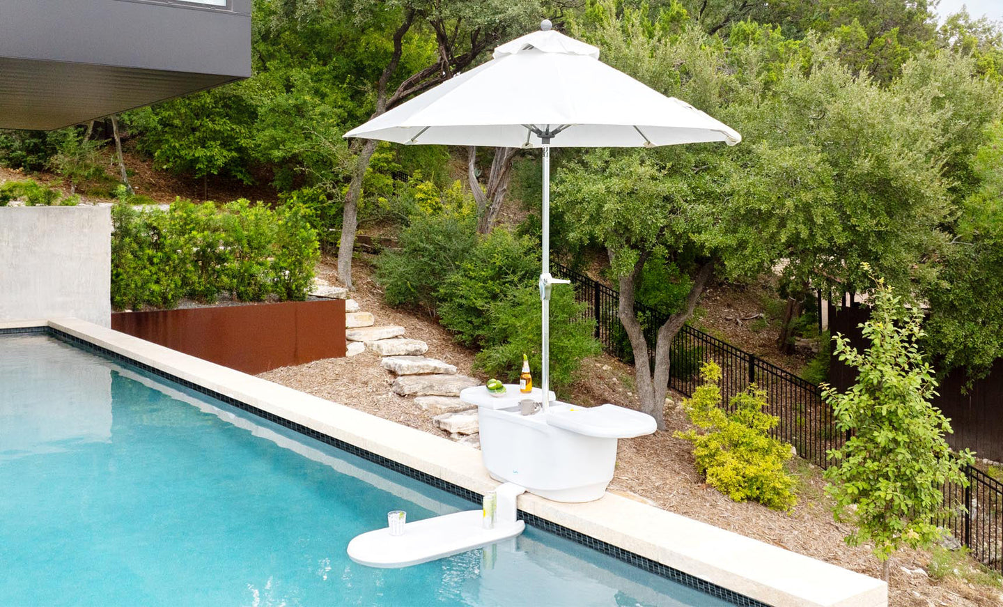 The PartyPad Swim-Up Bar by Ledge Lounger, with a white umbrella, is in the pool offering drinks and snacks. Surrounded by lush greenery, the serene outdoor area features stone and metal fencing, enhancing a tranquil atmosphere.