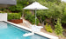 The PartyPad Swim-Up Bar by Ledge Lounger, with a white umbrella, is in the pool offering drinks and snacks. Surrounded by lush greenery, the serene outdoor area features stone and metal fencing, enhancing a tranquil atmosphere.