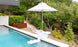 The PartyPad Swim-Up Bar by Ledge Lounger, with a white umbrella, is in the pool offering drinks and snacks. Surrounded by lush greenery, the serene outdoor area features stone and metal fencing, enhancing a tranquil atmosphere.