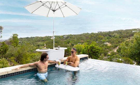 Two men relax in an infinity pool on submerged benches, using the PartyPad Swim-Up Bar by Ledge Lounger as a beverage center with drinks and a large umbrella. The pool offers scenic views of trees and hills under a clear sky.