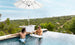 Two men relax in an infinity pool on submerged benches, using the PartyPad Swim-Up Bar by Ledge Lounger as a beverage center with drinks and a large umbrella. The pool offers scenic views of trees and hills under a clear sky.