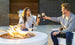 A man and woman, wearing casual sweaters and seated on outdoor chairs, smile and clink wine glasses in front of a lit Halo Aglow Bundle by Ledge Lounger. A grill is visible in the background on the patio.