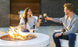 A man and woman, wearing casual sweaters and seated on outdoor chairs, smile and clink wine glasses in front of a lit Halo Aglow Bundle by Ledge Lounger. A grill is visible in the background on the patio.