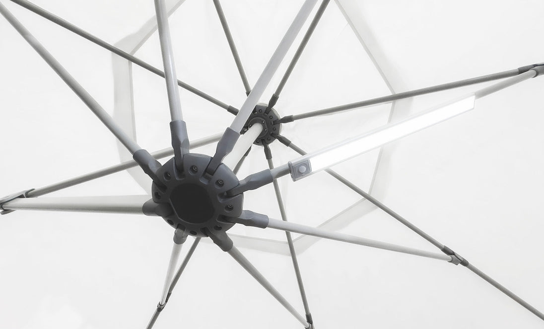 Close-up of the underside of a white umbrella with metal spokes radiating from a central hub. A Ledge Lounger Umbrella Light is attached to one spoke, creating a soft glow against the matching fabric background, ideal for an inviting patio setting.