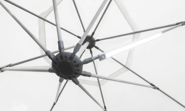 Close-up of the underside of a white umbrella with metal spokes radiating from a central hub. A Ledge Lounger Umbrella Light is attached to one spoke, creating a soft glow against the matching fabric background, ideal for an inviting patio setting.