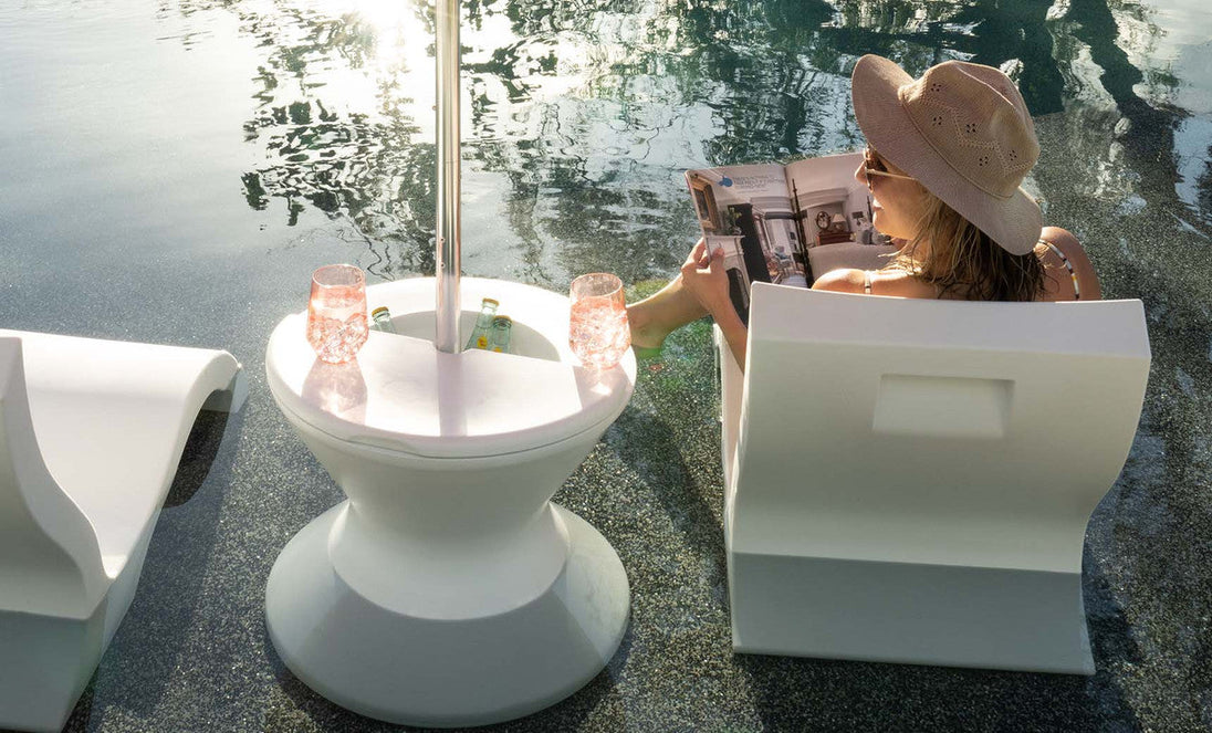 A woman relaxes outside, sitting partially submerged in a pool on a white chair with a wide-brimmed hat, reading a magazine. A round table with the Signature Side Table Lid by Ledge Lounger holds pink drinks under an umbrella reflecting light through its umbrella hole onto the water.