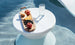 A Signature Side Table Lid with an umbrella hole by Ledge Lounger holds a wooden tray of sliced apples, orange segments, blackberries, and a peach in the pool. A glass of clear water sits beside it. The UV-resistant round white tables shadow adds to the serene and refreshing poolside atmosphere.